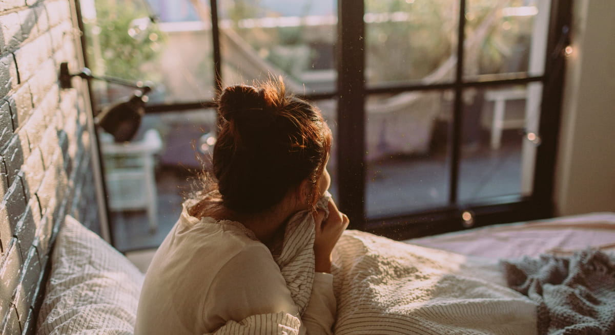Woman sitting on the edge of a bed in early morning light, looking out a window