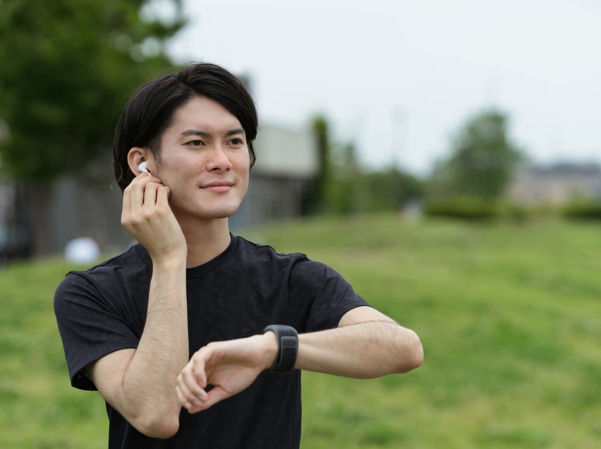 A young man in a black T-shirt standing on a grassy park lawn, glancing at the fitness watch on his wrist with a wireless earbud in one ear, checking his progress mid-walk