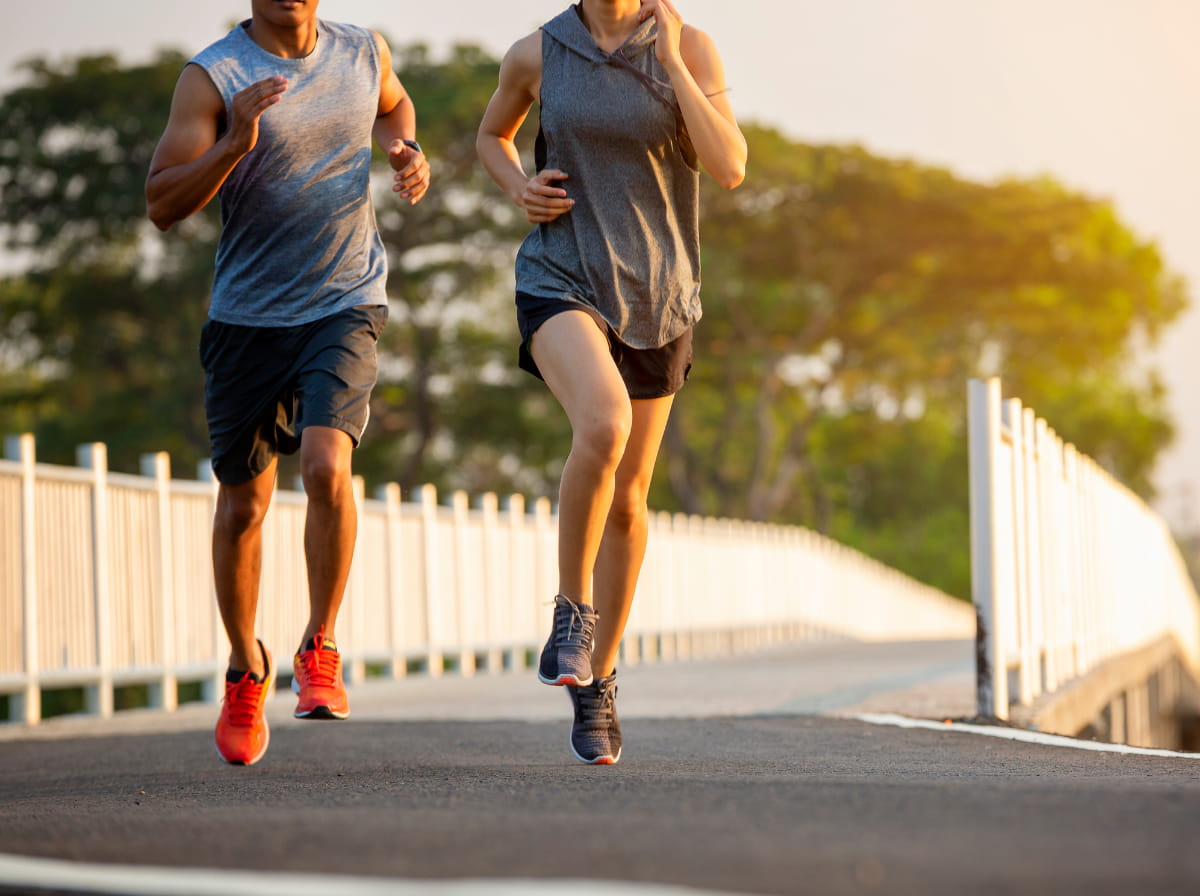 A man and woman jogging side by side across a railed footbridge at sunset, shown from the waist down in athletic wear and running shoes, moving at a brisk, purposeful pace